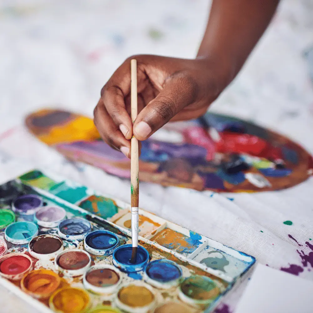 A hand holding a paintbrush over a watercolor palette, illustrating the hands-on art activities in the Therapeutic Nutritional Art Program.